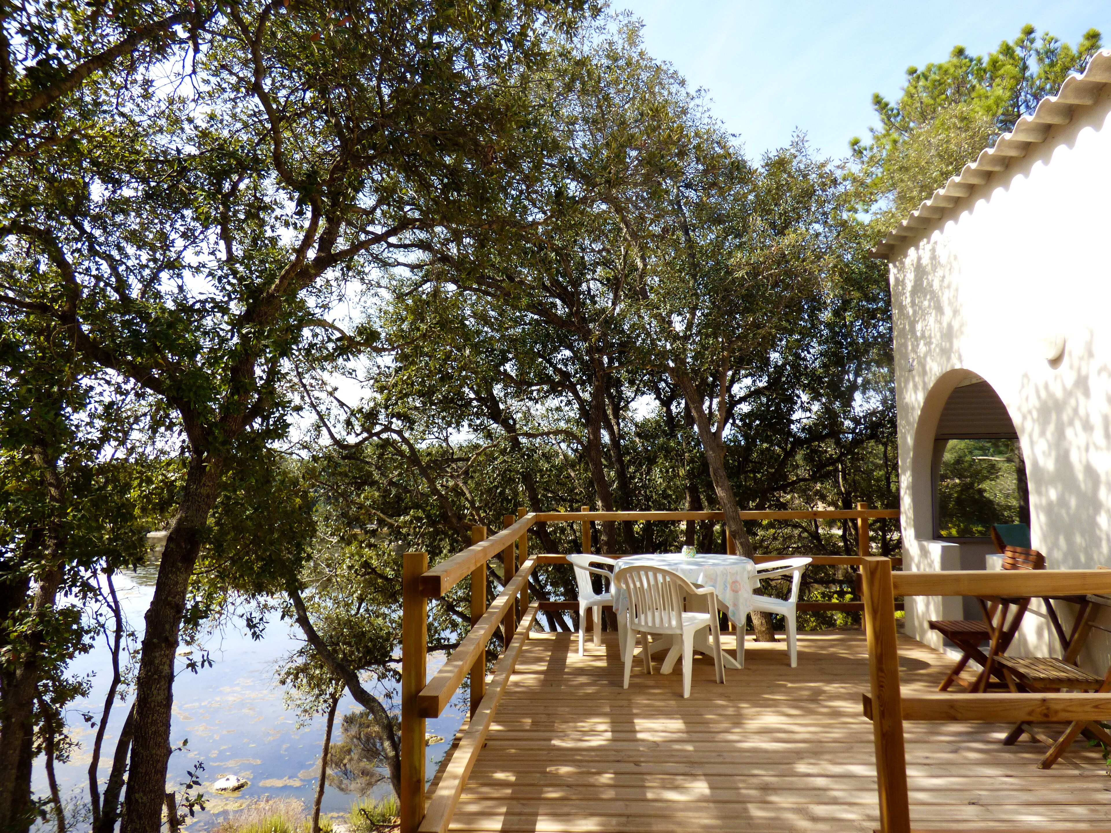 Eine Holzterrasse mit weißen Kunststoffstühlen und einem Tisch, mit Blick auf ein Gewässer, umgeben von Bäumen.
