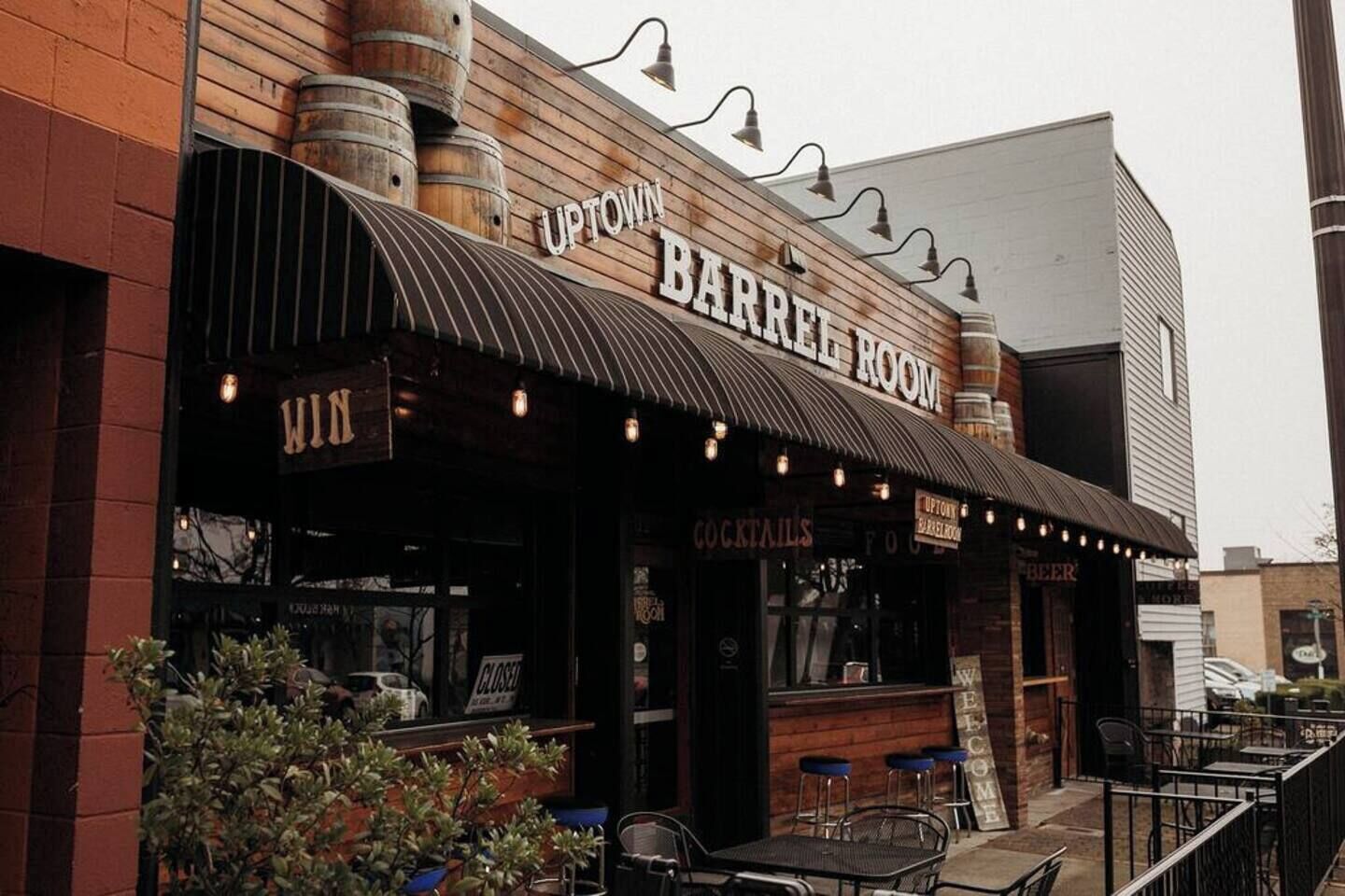 A bar with a rustic wooden facade, outdoor seating, and a sign reading "UPTOWN BARREL ROOM".