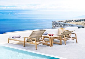 Ein Poolbereich mit zwei Holzliegestühlen, einem kleinen Tisch und einer Steinmauer mit Blick auf das Meer.