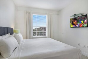 A hotel room with a bed, curtains, a window with blinds, a television on the wall, and a power outlet.