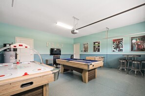 A pool table, a foosball table, and a bar area with stools in a room with teal walls and a ceiling-mounted projector.