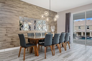 Un comedor con una gran mesa de madera y seis sillas, una lámpara moderna y un ventanal con vistas a una piscina y casas.