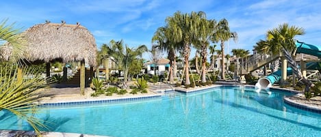 A swimming pool with a slide, surrounded by palm trees and a thatched-roof structure.