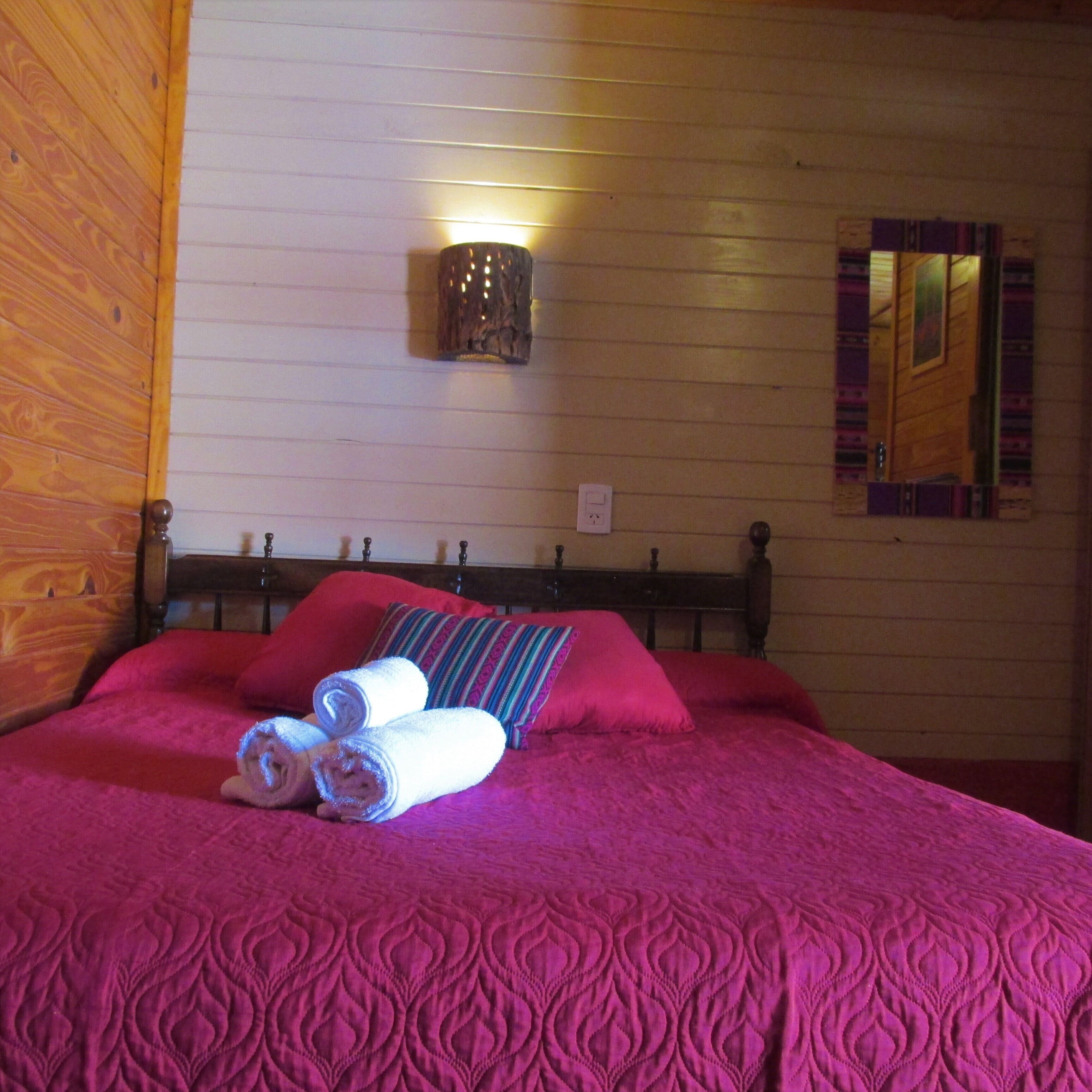 A bedroom with a wooden headboard, a bed with a pink quilt, rolled towels, a wall-mounted light, and a mirror.