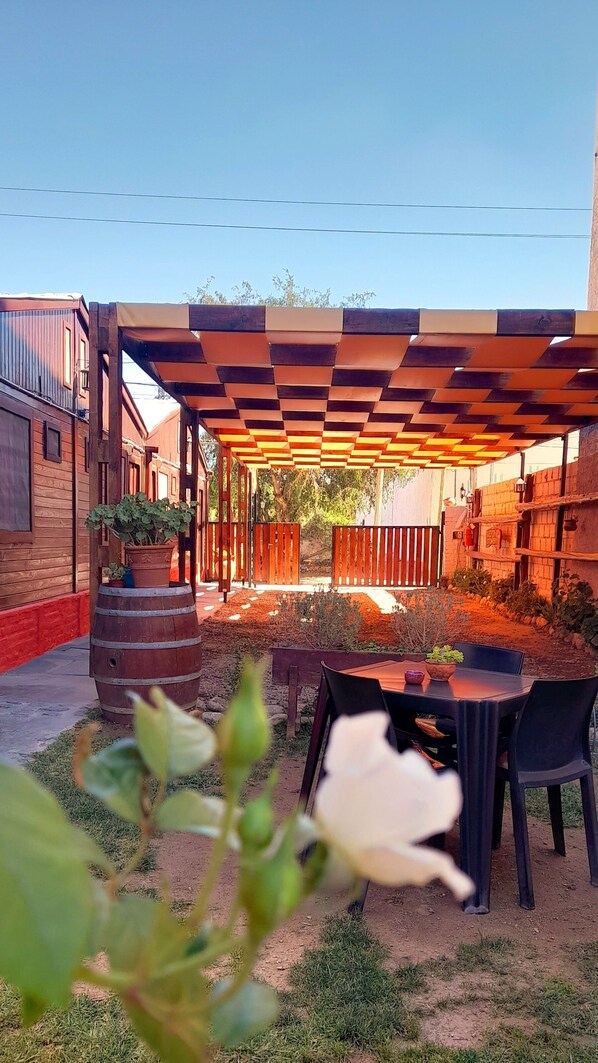 Un patio al aire libre con una pérgola de madera, una mesa con sillas y plantas en macetas.