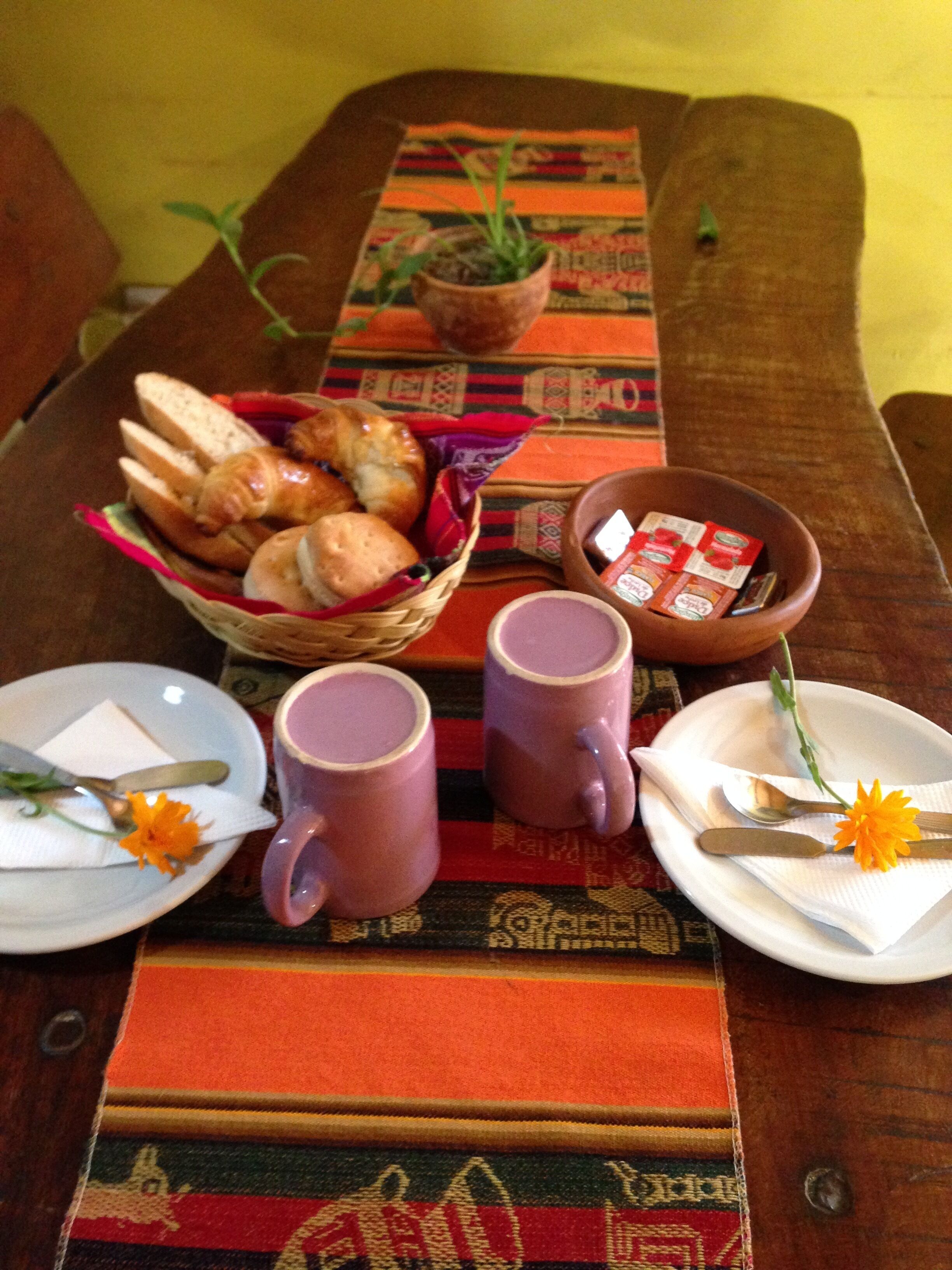 Une table à manger avec un set de table en fibres tressées, un panier de pain, deux tasses, une assiette décorée d’une fleur et un bol de thé.
