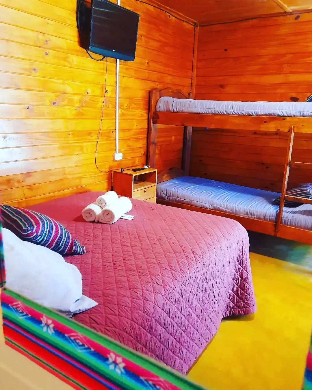 Quarto com beliche, coberto com edredom rosa acolchoado, uma mesinha de cabeceira de madeira e televisão fixada na parede.