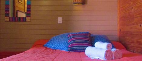 A bedroom with a wooden wall, a bed with pink bedding, two pillows, and a wall-mounted lamp.