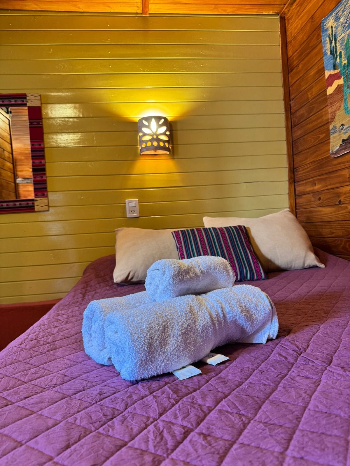 A bed with a purple quilt, two rolled towels, and a wall-mounted light fixture.