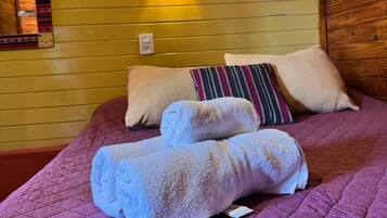 A bed with a purple quilt, two rolled towels, and a wall-mounted light fixture.