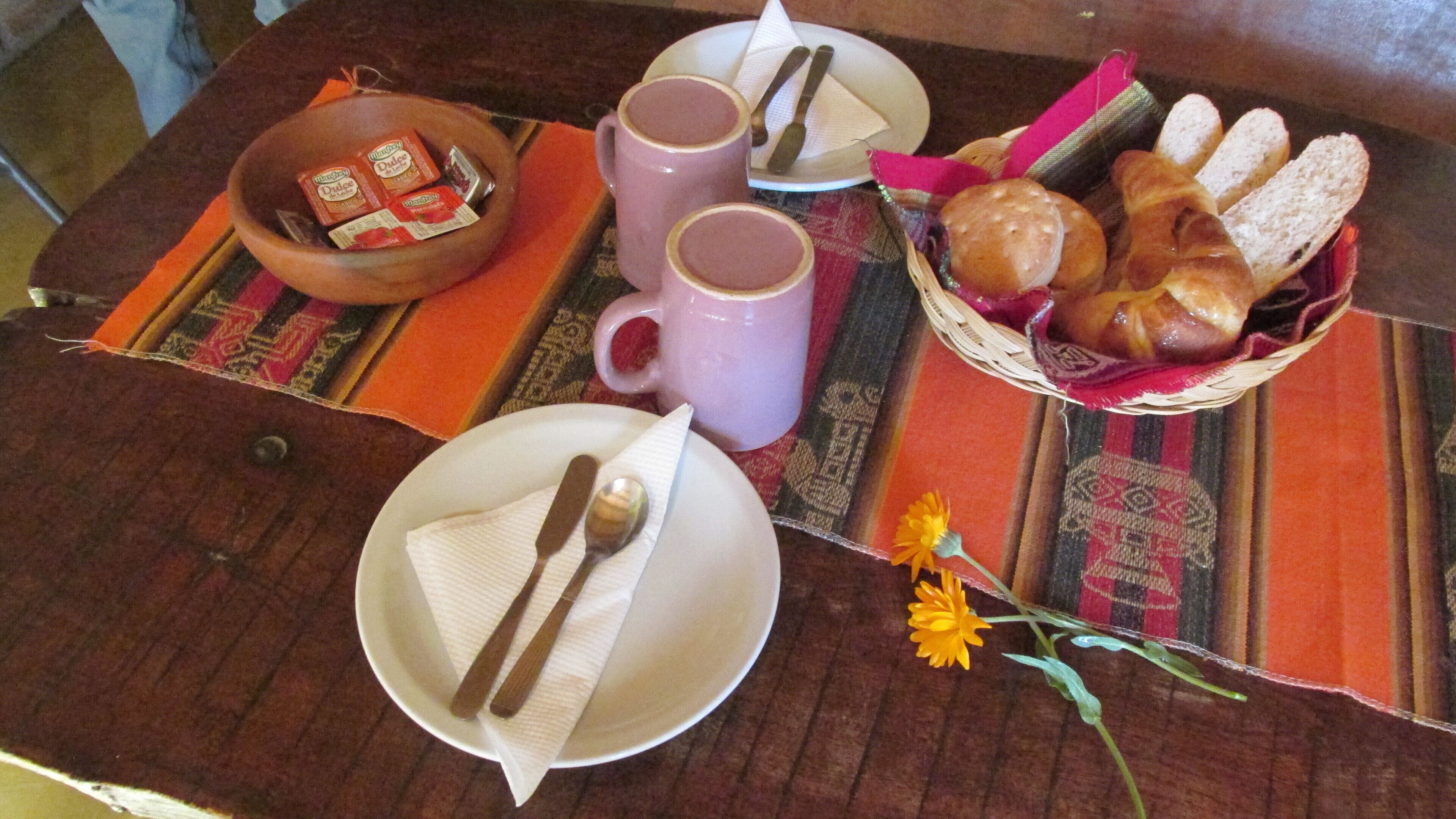 Une table en bois avec une assiette, deux tasses, un panier de pain et un petit bol de collations.