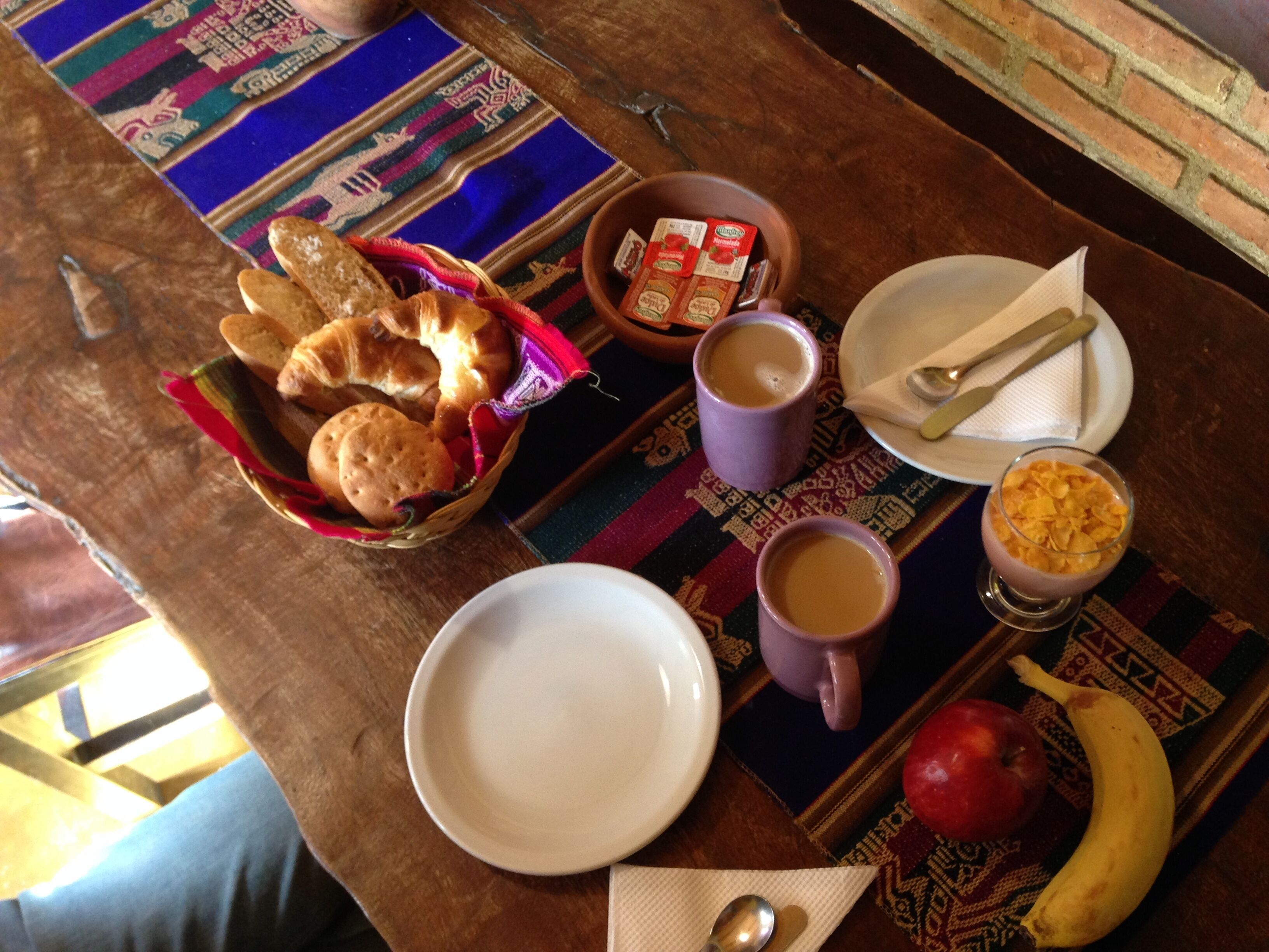 Una mesa de desayuno con pan, pasteles, un tazón de cereal, dos tazas de café, un plato con una cuchara y un plátano.
