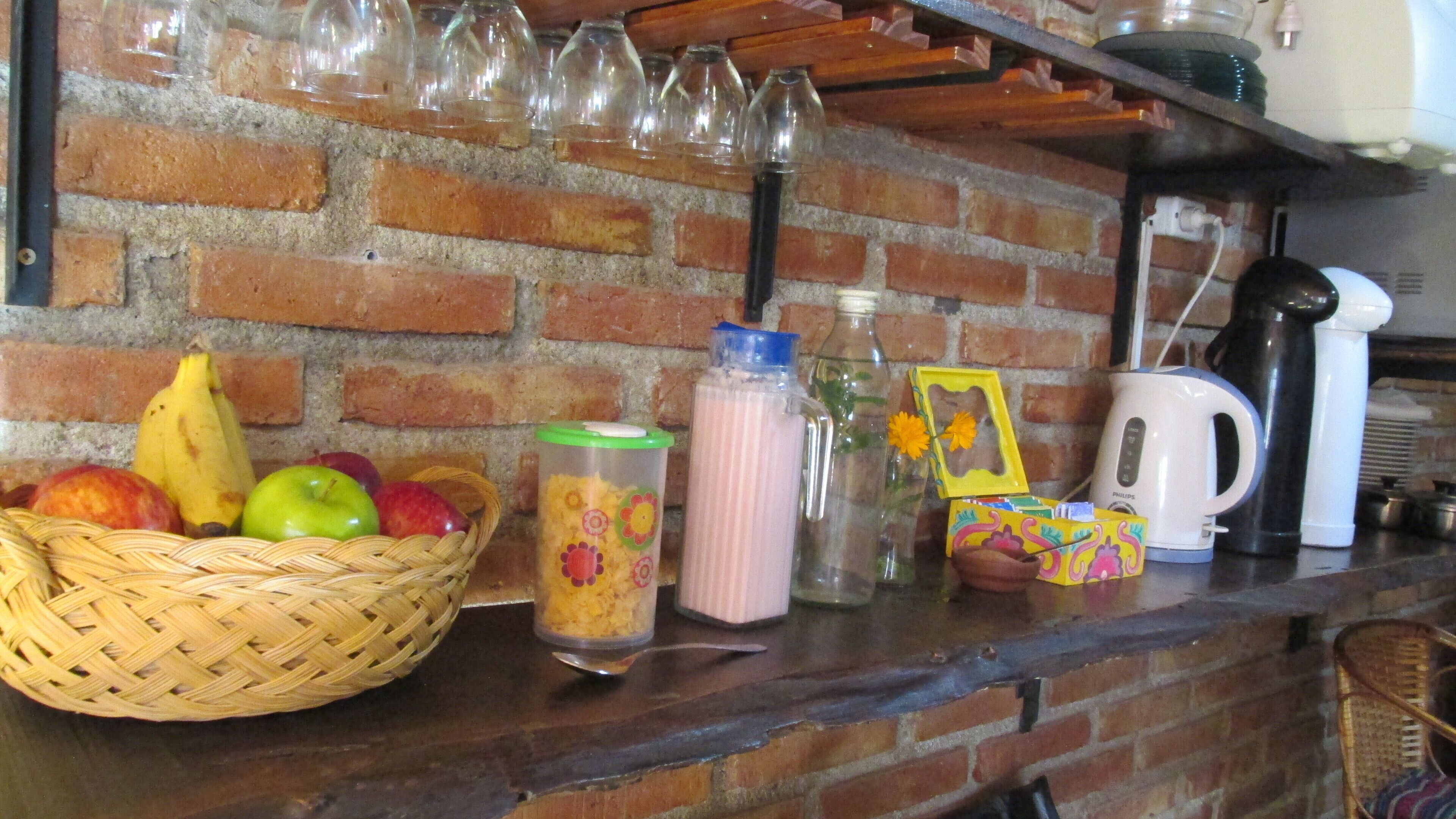 A kitchen counter with a brick wall, a wicker basket of fruits, a glass container with a floral design, a pink bottle, a yellow box with a floral design, and a white coffee maker.