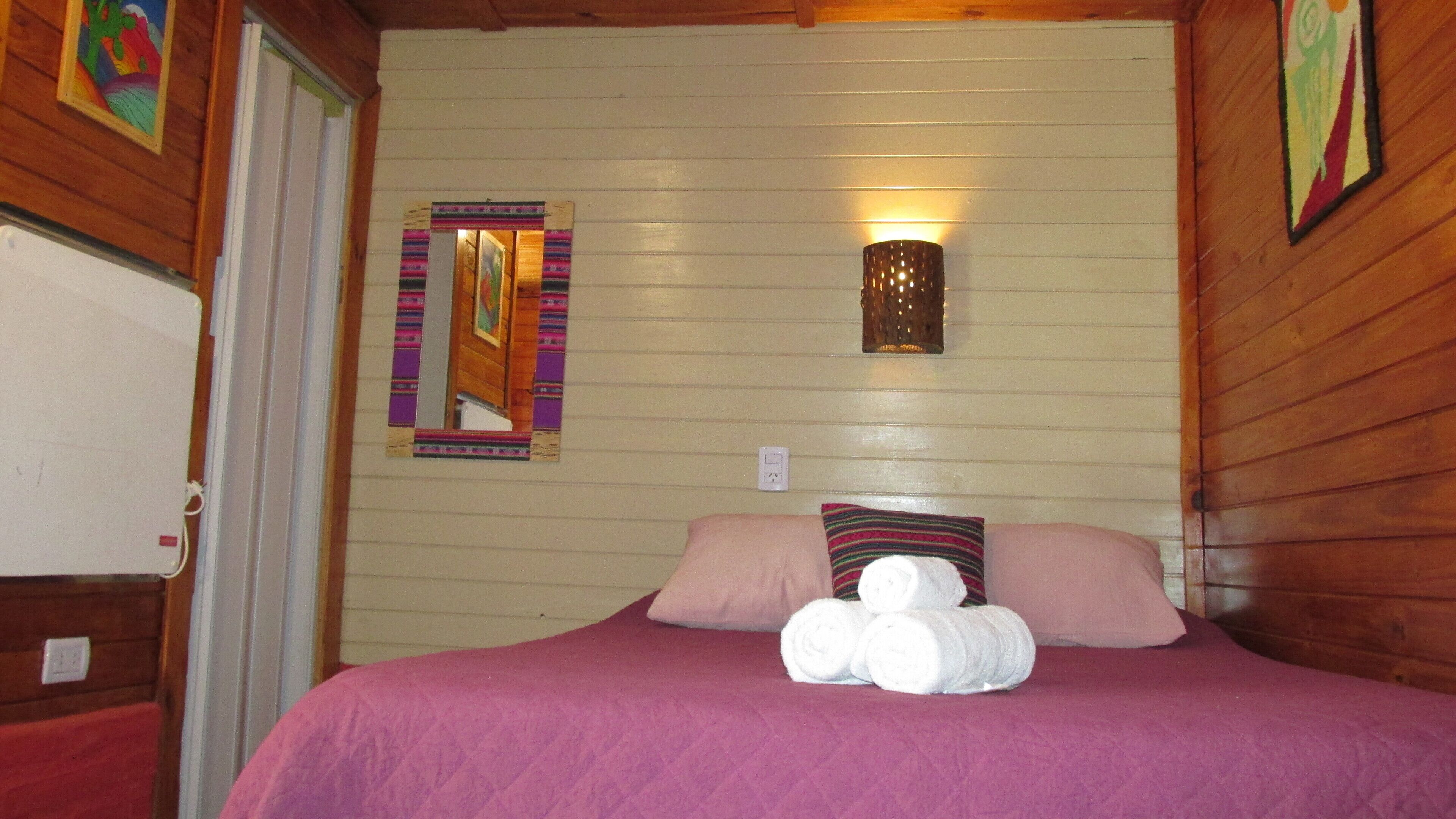 Una habitación con paredes de madera, una cama con cubrecama rosa, dos almohadas y una lámpara fijada a la pared.