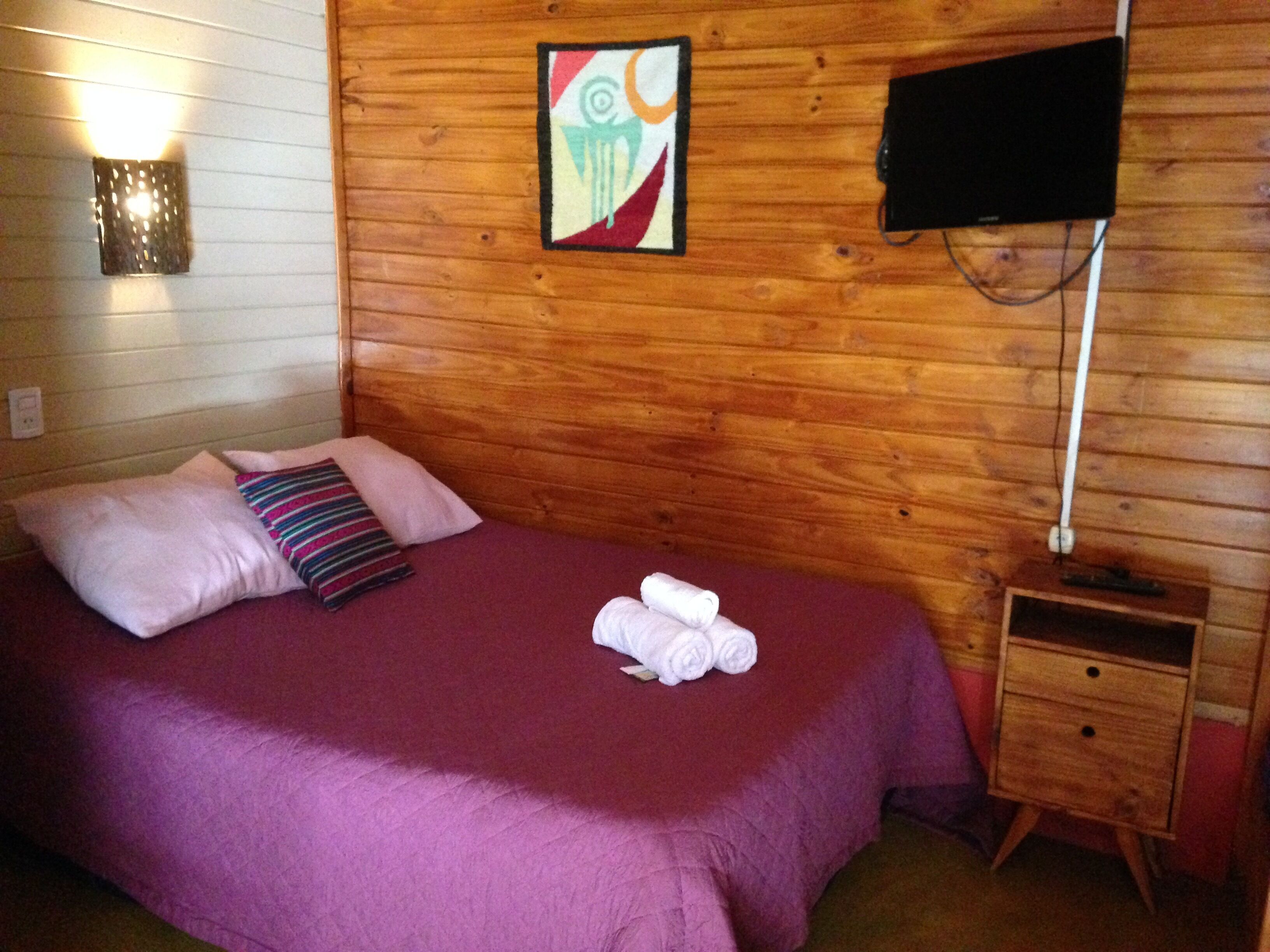 A room with a wooden wall, a bed with a purple bedspread, a TV on a stand, and a small wooden nightstand.