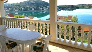 A balcony with a table and chairs, overlooking a lake with buildings and hills in the background.