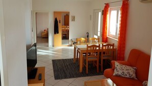 A living room with a sofa, a dining table with chairs, a TV, and a kitchen area in the background.