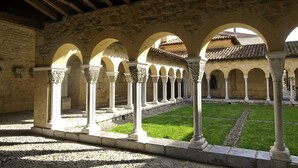 A courtyard with stone arches and columns, a grassy area, and a building with a tiled roof in the background. - Paradise Chalets and Camping (Sarrecave)