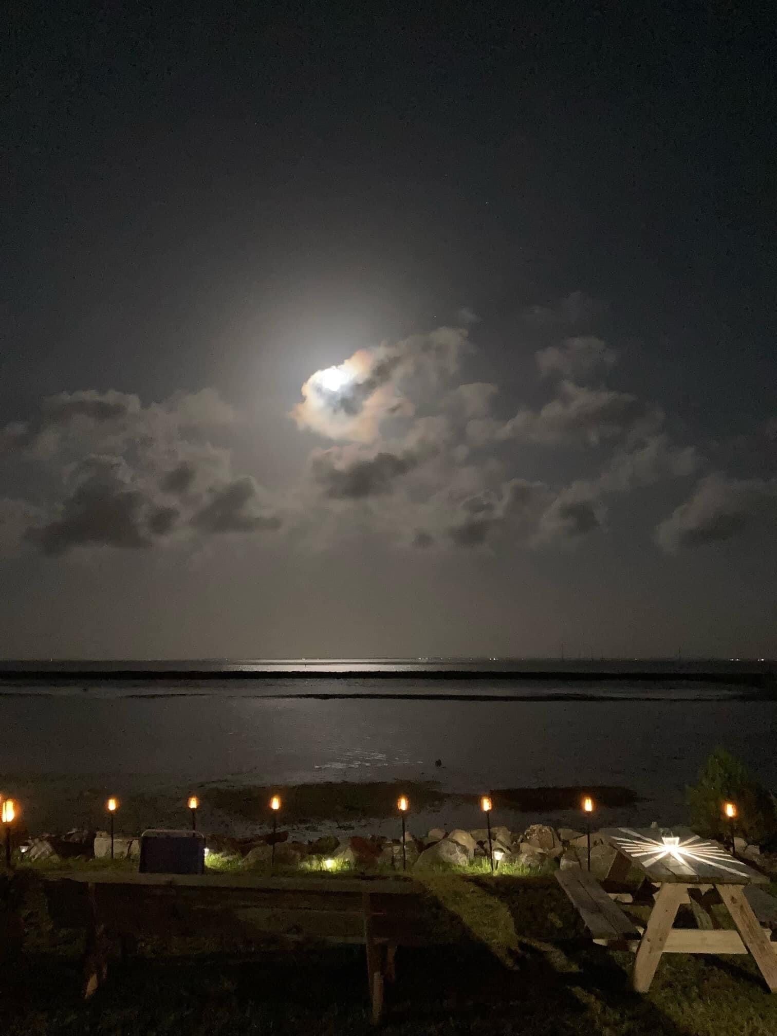 Cena noturna de praia com lua cheia, nuvens e uma fileira de velas acesas na margem.