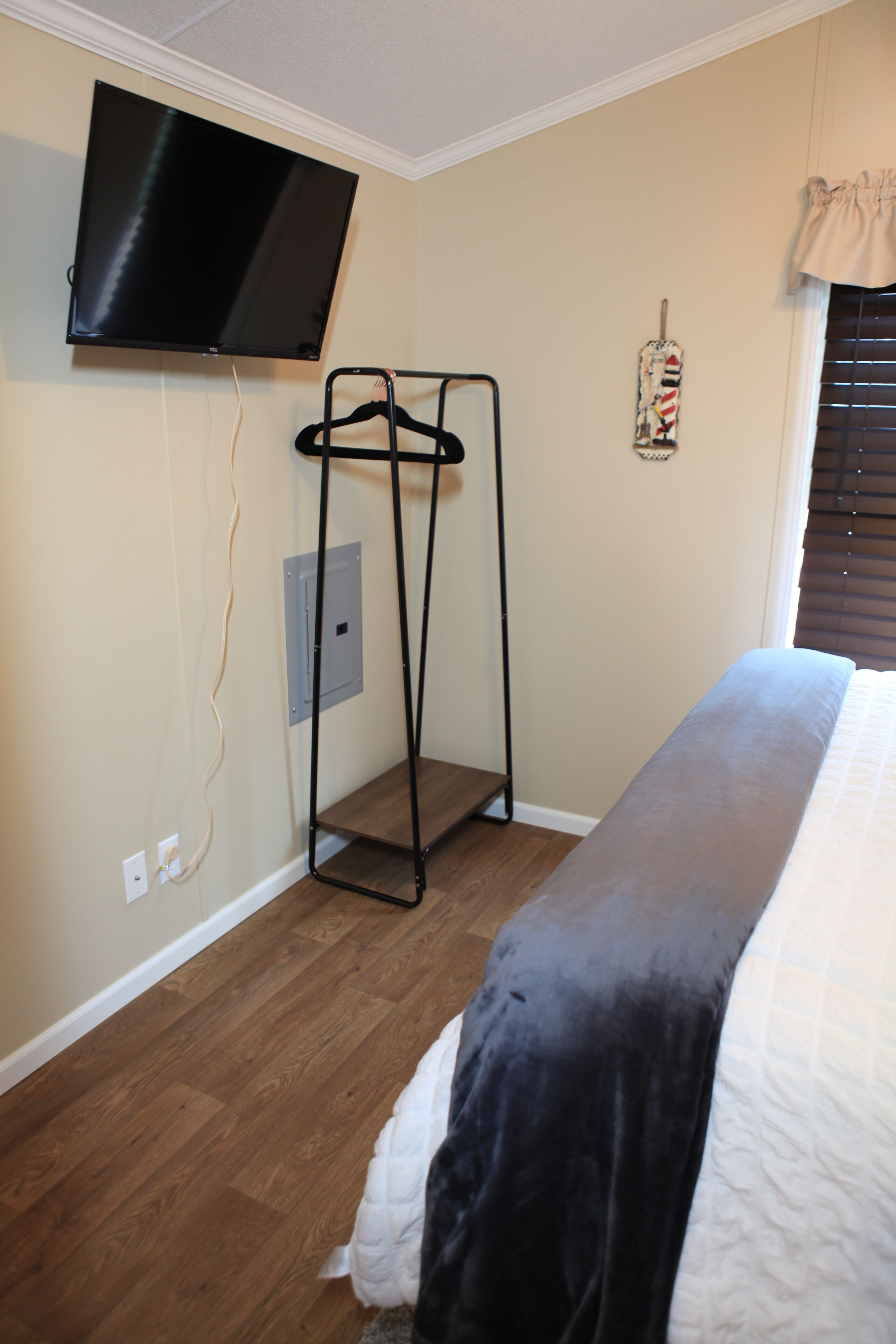 Un dormitorio con cama, televisor montado en la pared, perchero y ventana con persianas.