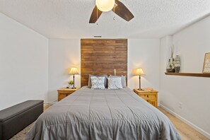 Un dormitorio con cabecera de madera, dos mesitas de noche con lámparas, una colcha gris y un ventilador de techo.