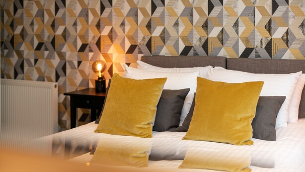 A neatly made bed with a patterned wall in the background. - The Lucas - Donnini Apartments (Ayr)