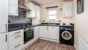 A compact kitchen with white cabinets, a black countertop, a built-in oven, a washing machine, and a window with a view. - The Lucas - Donnini Apartments (Ayr)
