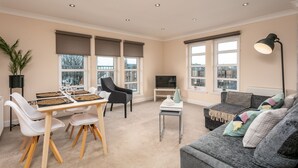 A modern living room with a dining area, large windows, a flat-screen TV, and a cozy seating area with cushions. - The Lucas - Donnini Apartments (Ayr)
