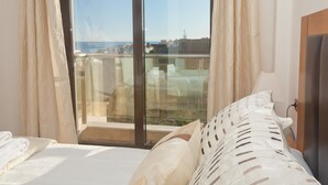 Une chambre avec une grande fenêtre, un lit avec une literie blanche et une vue sur la mer et les bâtiments.