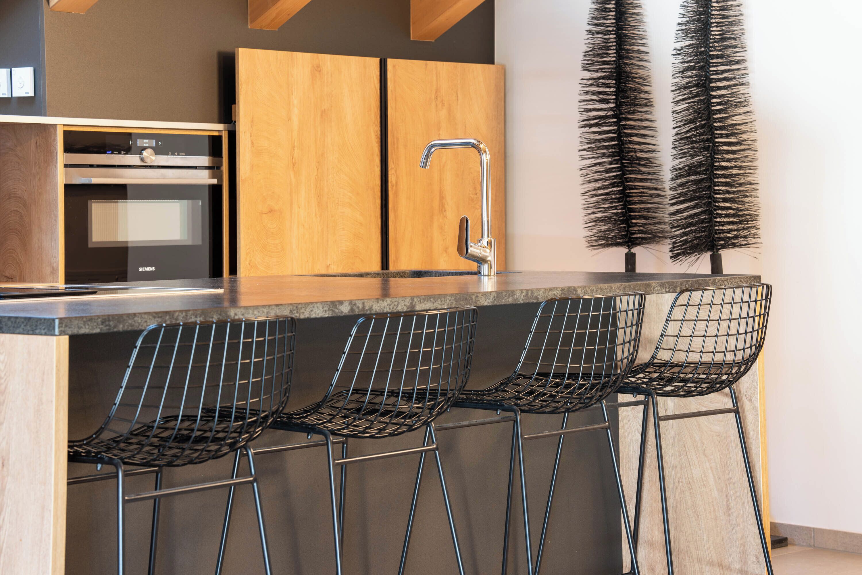A kitchen with a built-in oven, a wooden cabinet, a stainless steel faucet, and three wire mesh bar stools.