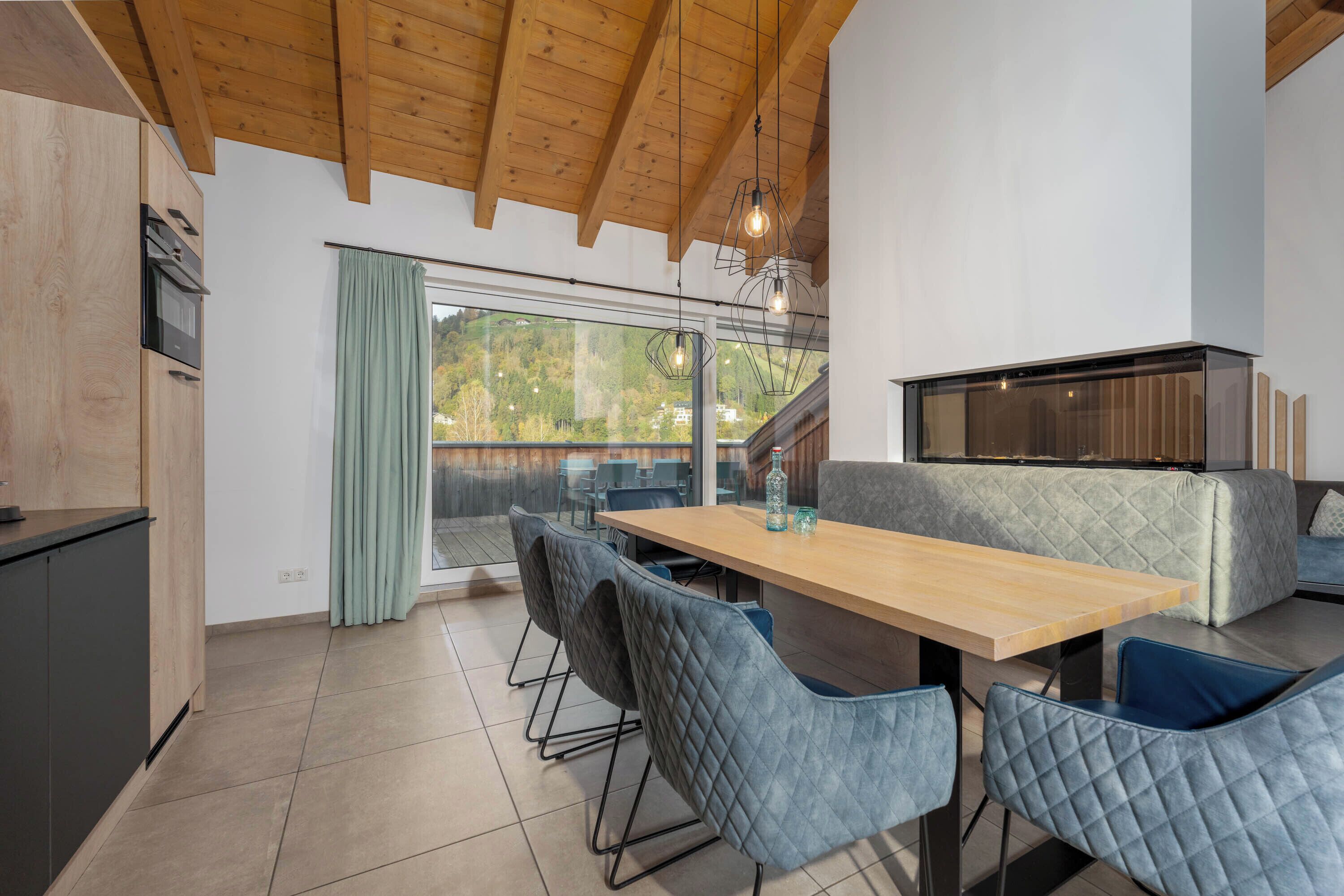 Una cocina moderna con vigas de madera, un amplio ventanal con cortinas, un comedor con mesa de madera y sillas azules, y un banco empotrado con respaldo acolchado.