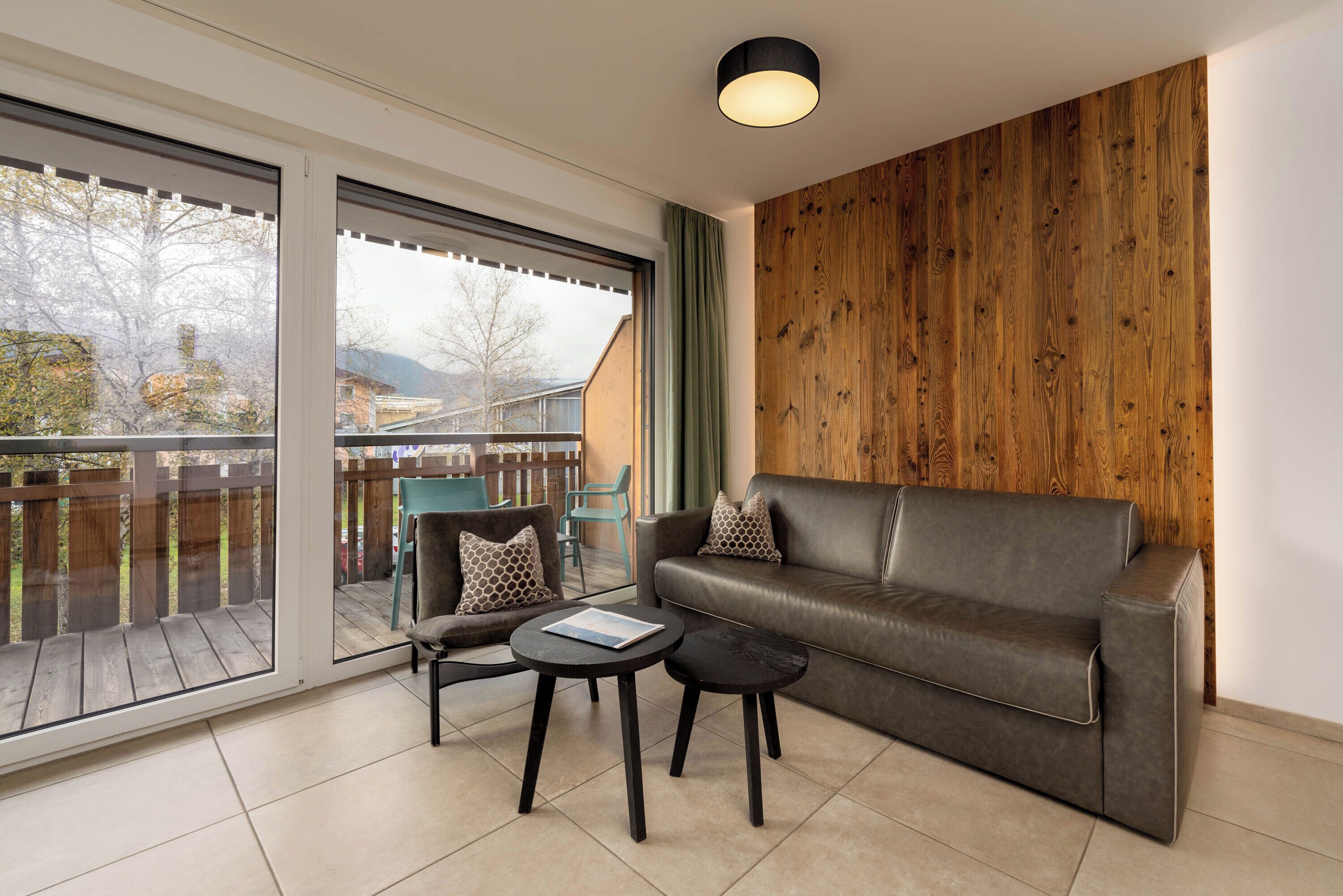 Un salon moderne avec un canapé en cuir, deux tables noires et une grande porte-fenêtre coulissante donnant sur une terrasse extérieure.
