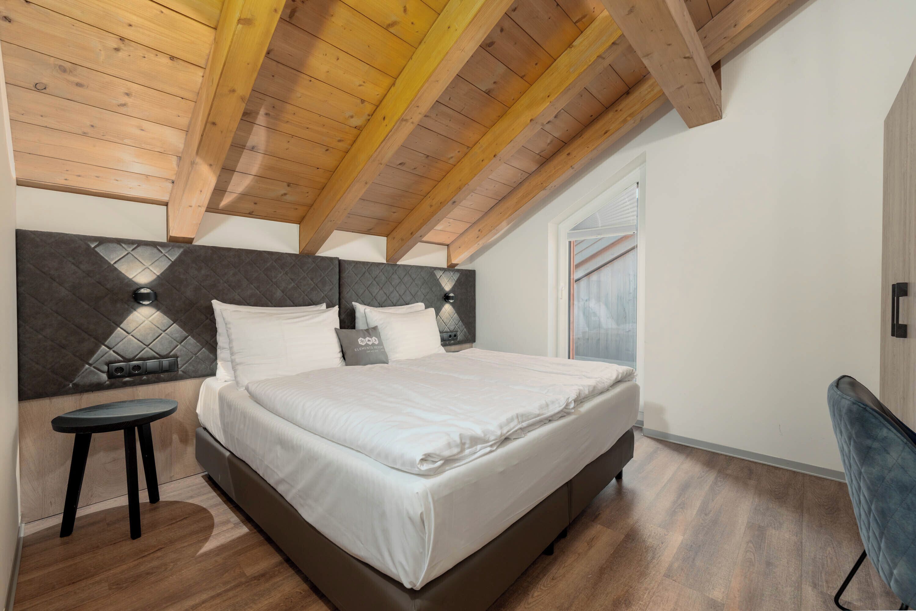 A bedroom with a bed, a small stool, a window, and wooden ceiling beams.
