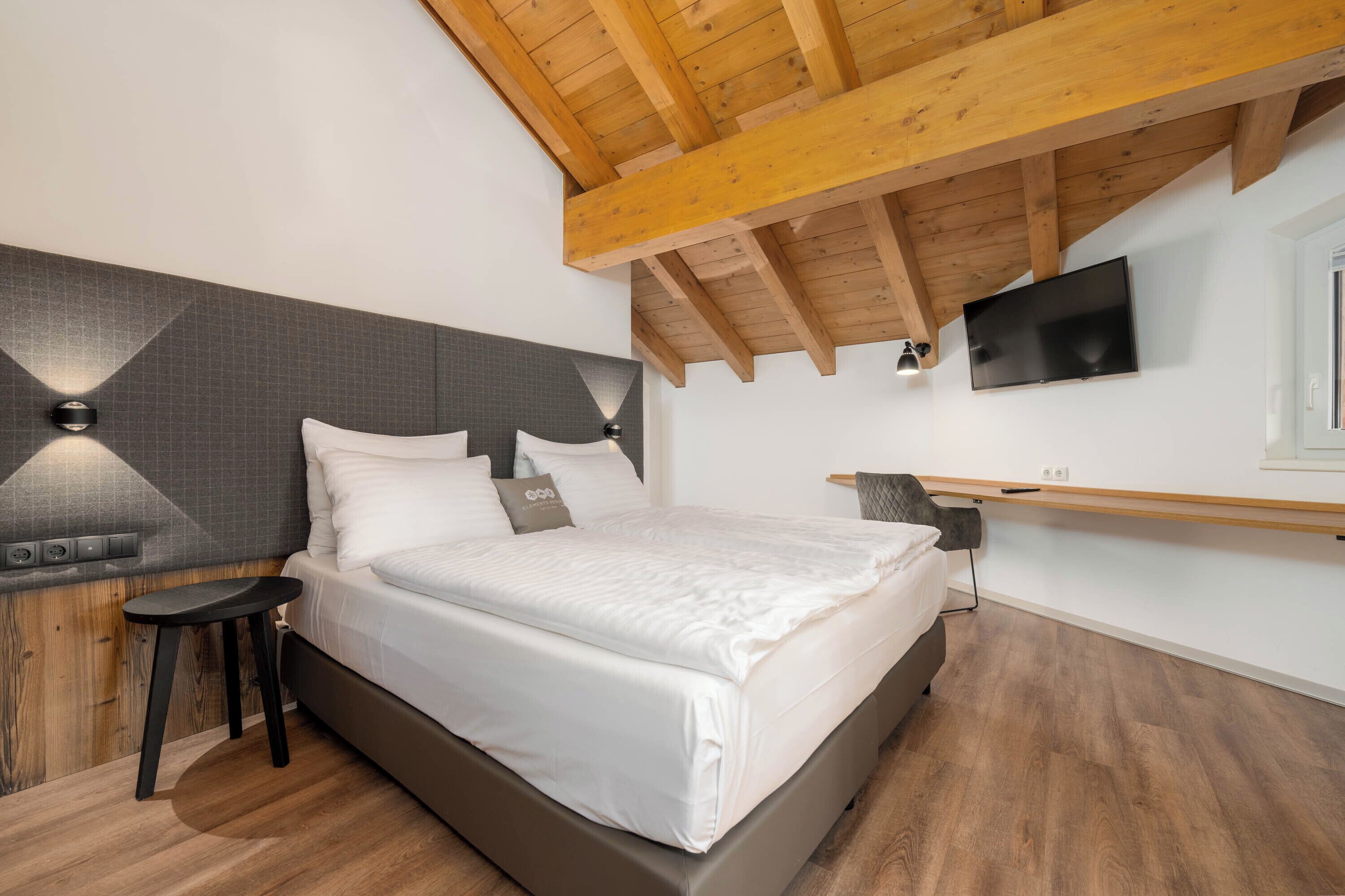 A bedroom with a bed, a nightstand, a TV mounted on the wall, and a wooden ceiling.