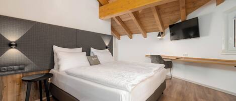 A bedroom with a bed, a nightstand, a TV mounted on the wall, and a wooden ceiling.