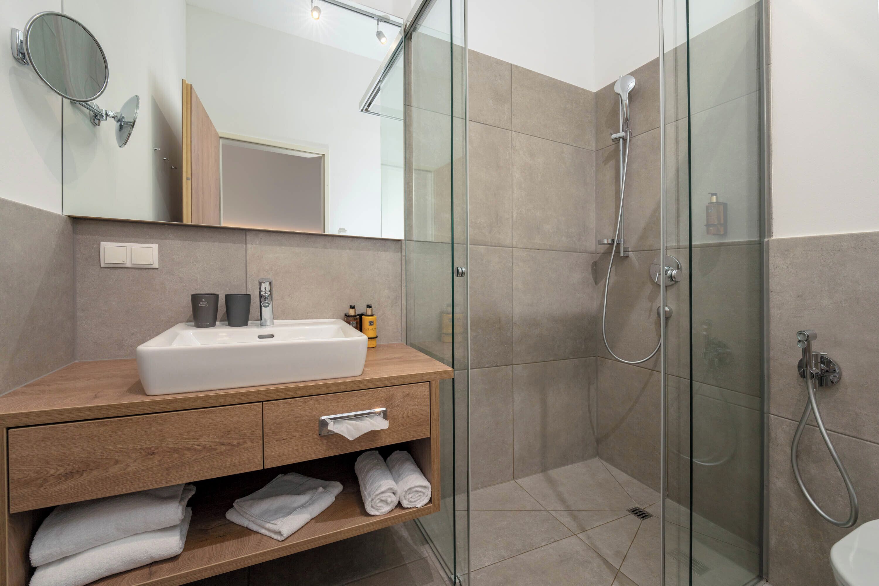 Un baño moderno con ducha envuelta en cristal, tocador de madera con lavamanos y un espejo.