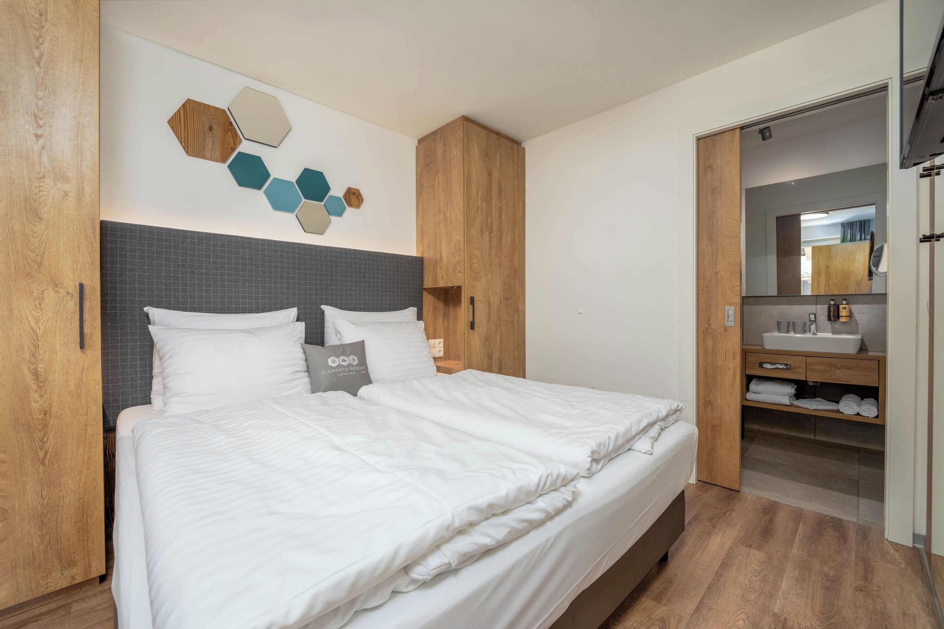 Une chambre moderne avec un grand lit, une armoire en bois et une salle de bain équipée d’un miroir et d’un lavabo.
