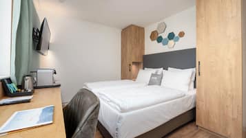 Quarto de hotel moderno com uma cama bem arrumada, uma escrivaninha com um laptop e uma cafeteira, e uma TV fixada na parede.