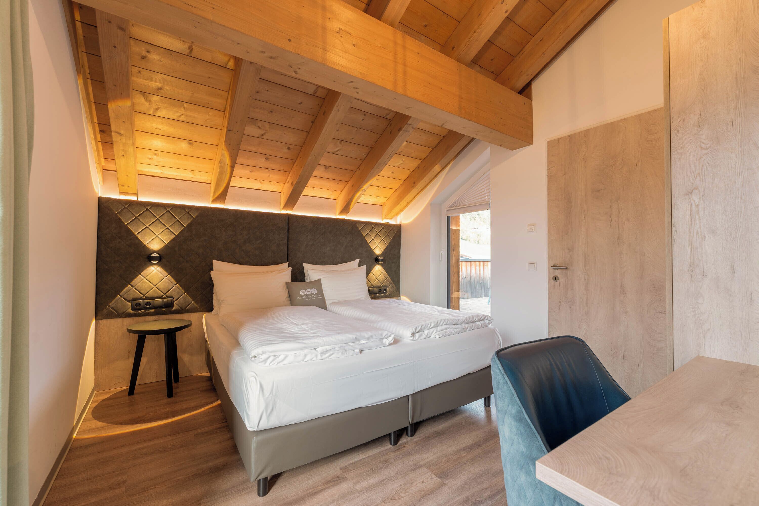 A bedroom with a wooden ceiling, a bed with white bedding, a small bedside table, and a chair.
