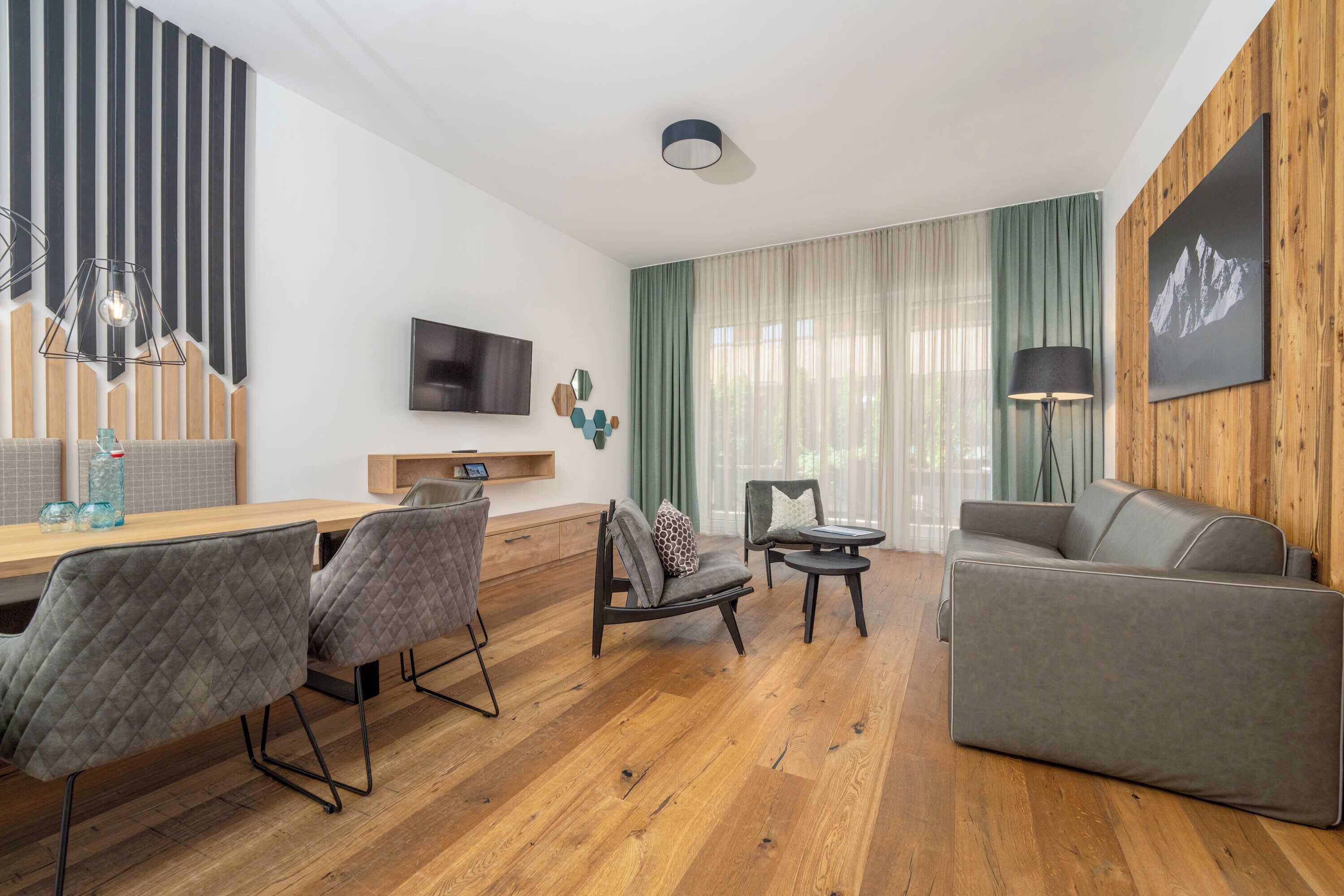 Een moderne woonkamer met een houten vloer, een grijze bank, een eettafel met stoelen, een aan de muur bevestigde televisie en een raam met gordijnen.