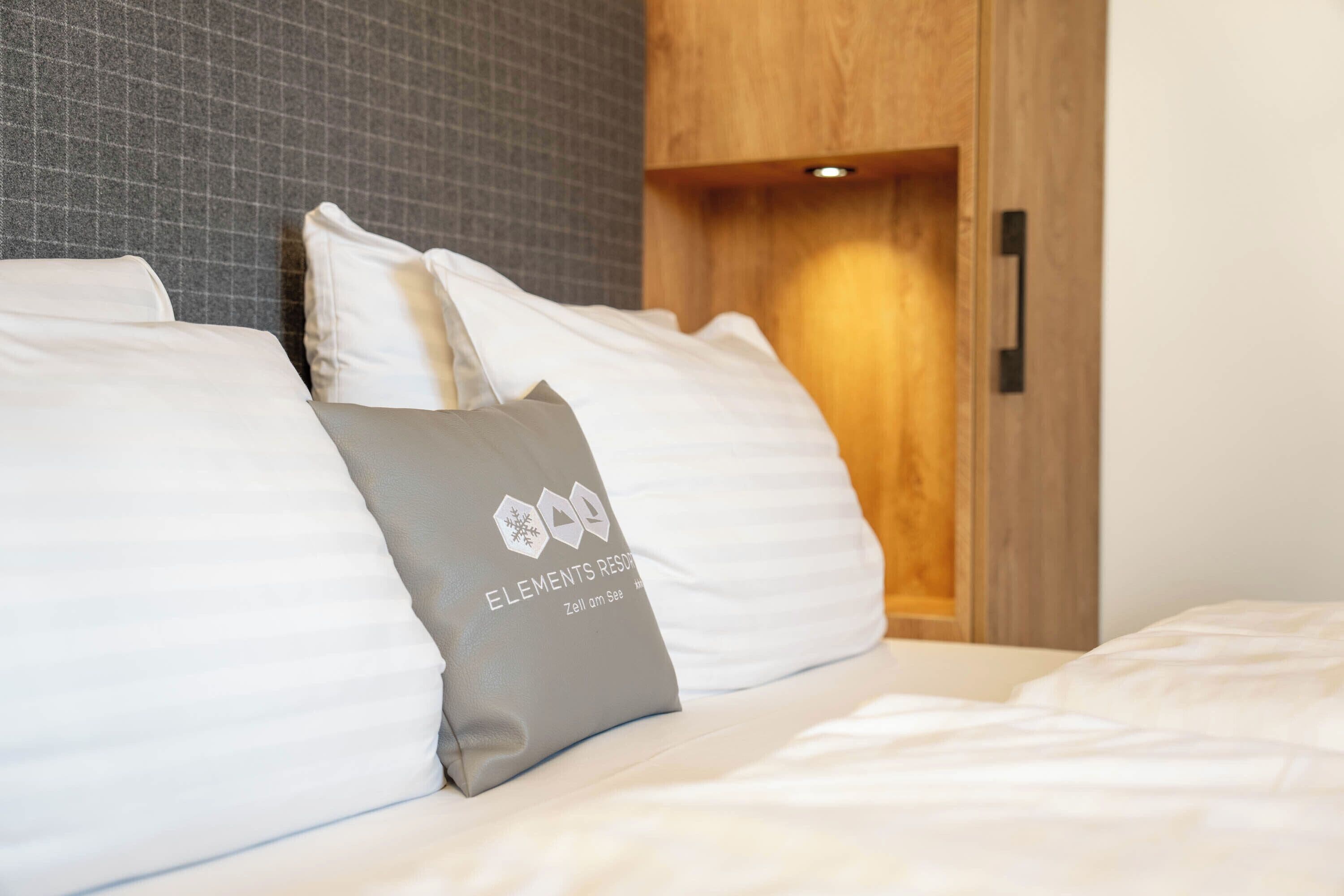 A neatly made bed with white bedding and a grey pillow with a logo.