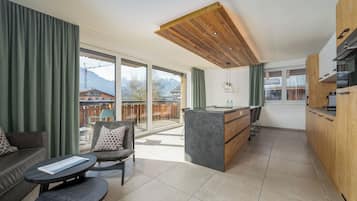 Una cocina moderna con armarios de madera y una isla central, un comedor con mesa y sillas, y un amplio ventanal corredero que da a un balcón con vistas a las montañas.