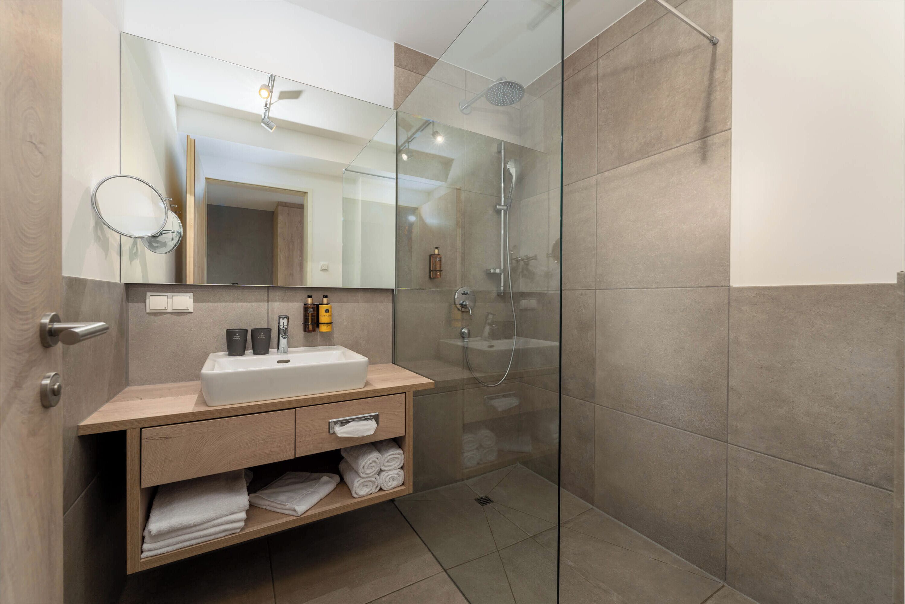 Une salle de bain moderne avec une douche à parois de verre, un grand miroir et un meuble-lavabo en bois doté d’un évier et d’un espace de rangement pour les serviettes.