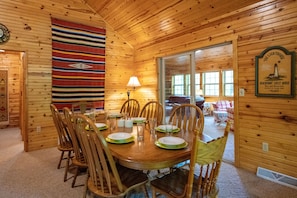 A wooden interior with a round dining table set for six, surrounded by wooden chairs. A colorful woven wall hanging is visible on the left, and a framed sign on the right wall indicates the property is a beachfront guest house.