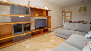 Ein modernes Wohnzimmer mit Flachbildfernseher, Holzregalen, einem grauen Sofa und einem Essbereich mit Tisch und Stühlen.