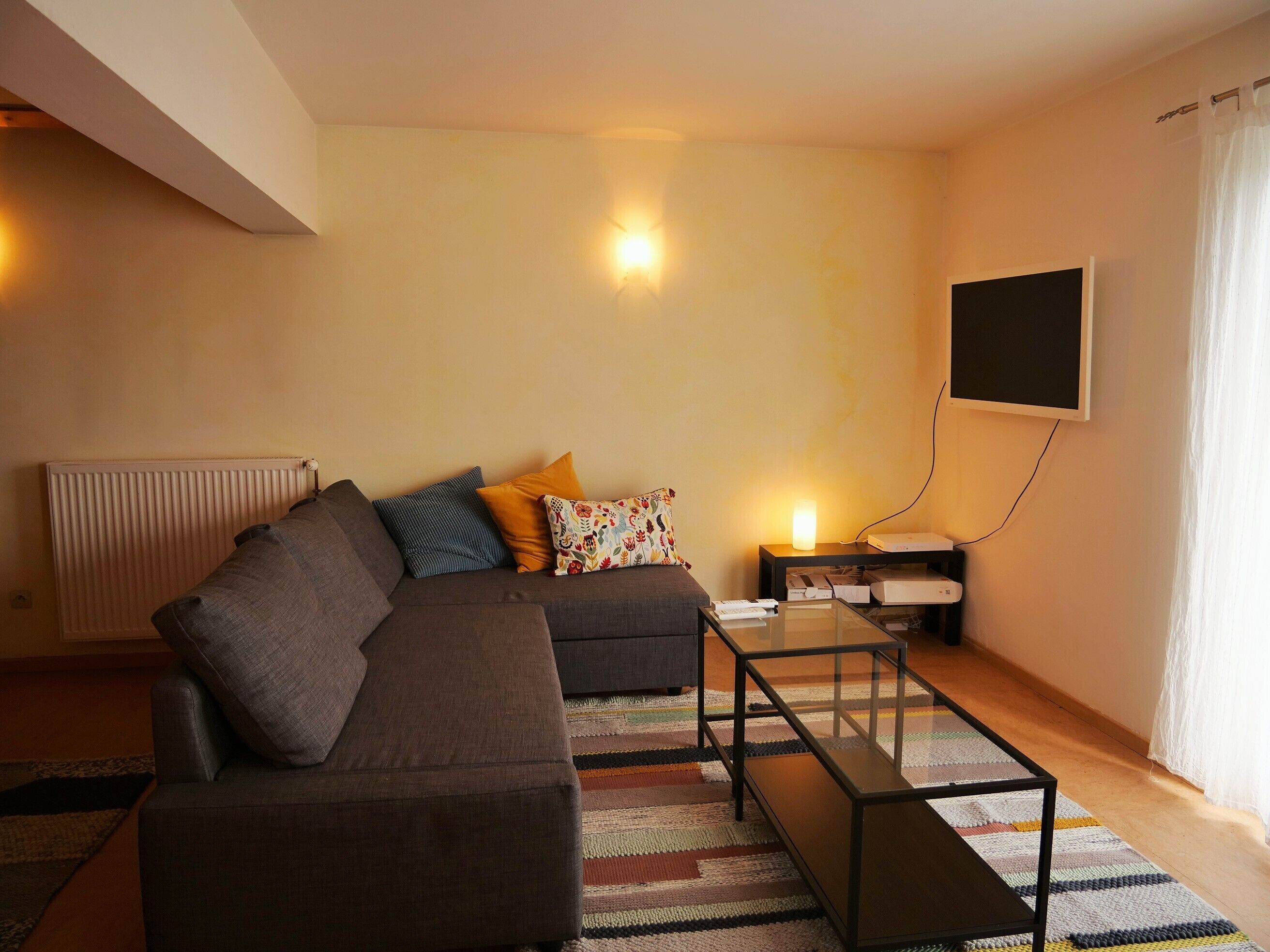 Uma sala de estar com sofá seccional, mesa de centro de vidro, tapete estampado, mesa lateral com abajur e televisão de tela plana na parede.