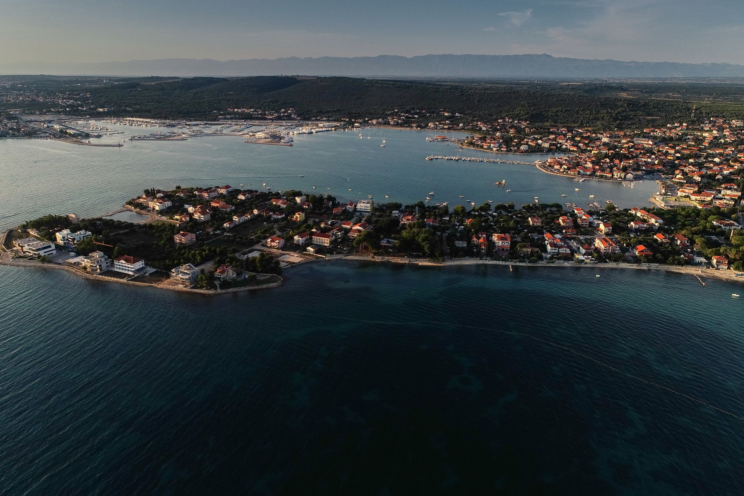 An aerial view of a coastal town with a marina, residential buildings, and a hilly landscape in the background.
