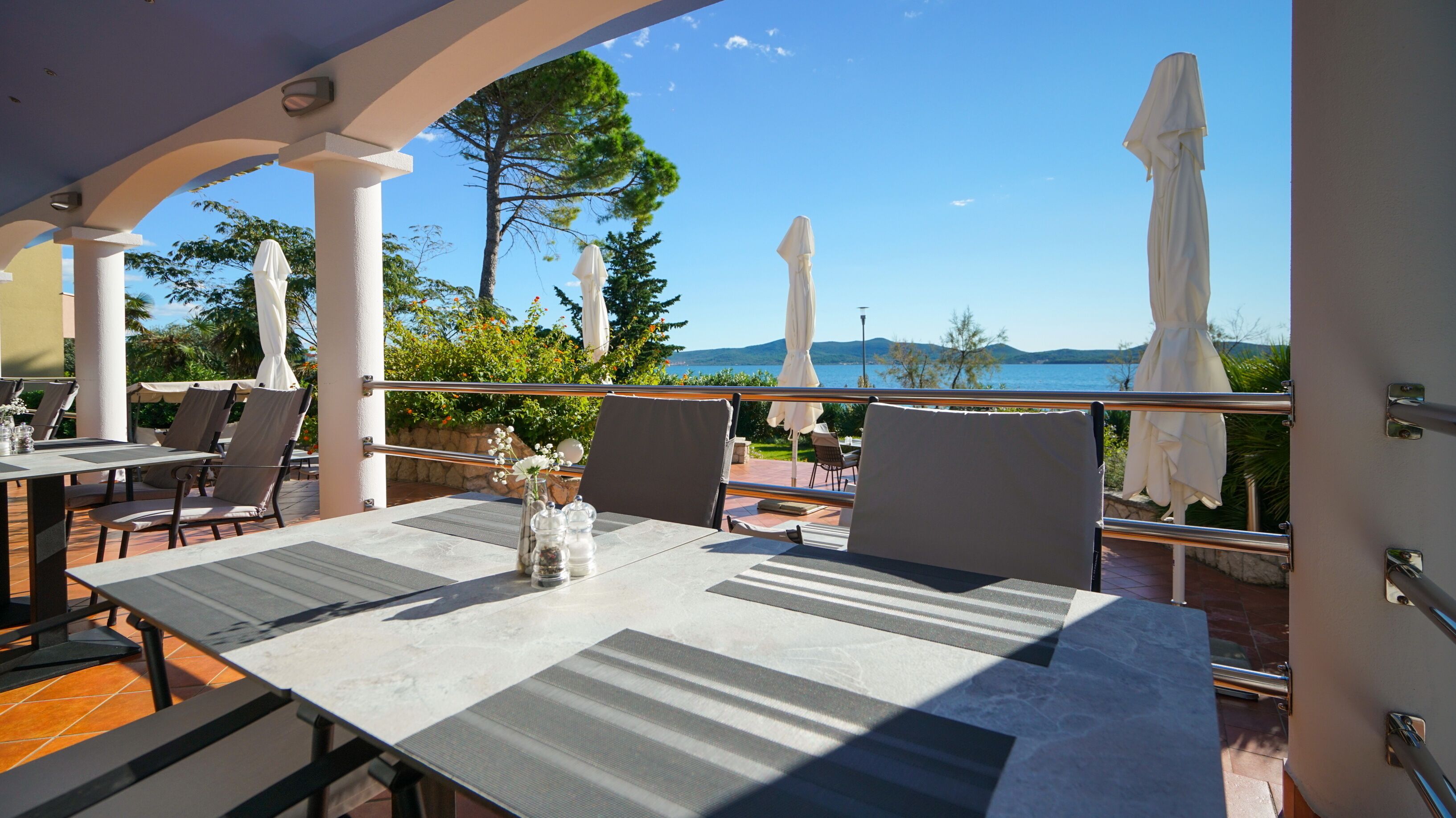 An outdoor dining area with tables and chairs, overlooking a scenic view of the sea and mountains.