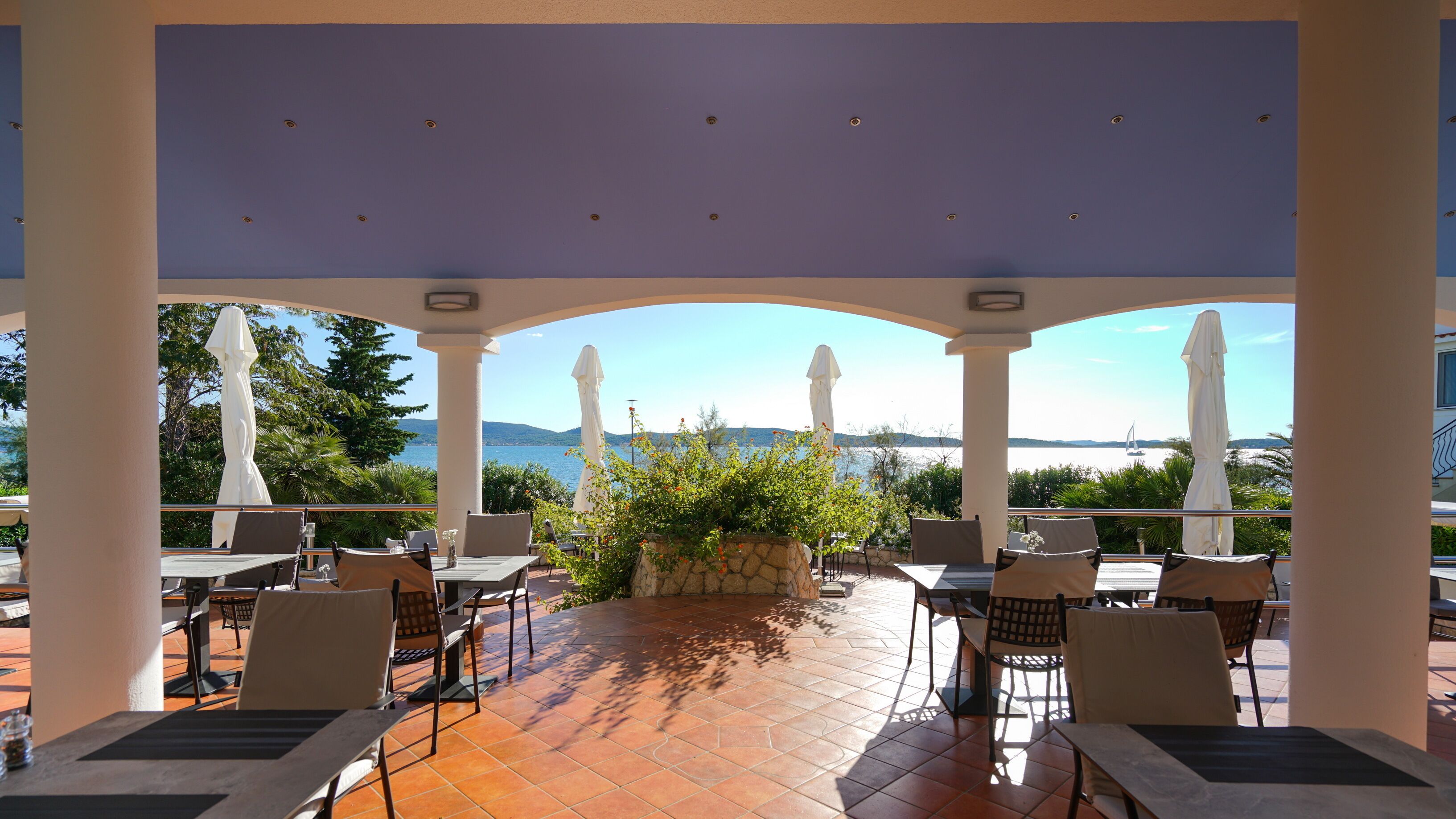 A covered outdoor seating area with tables and chairs, overlooking a scenic view of the sea and mountains.