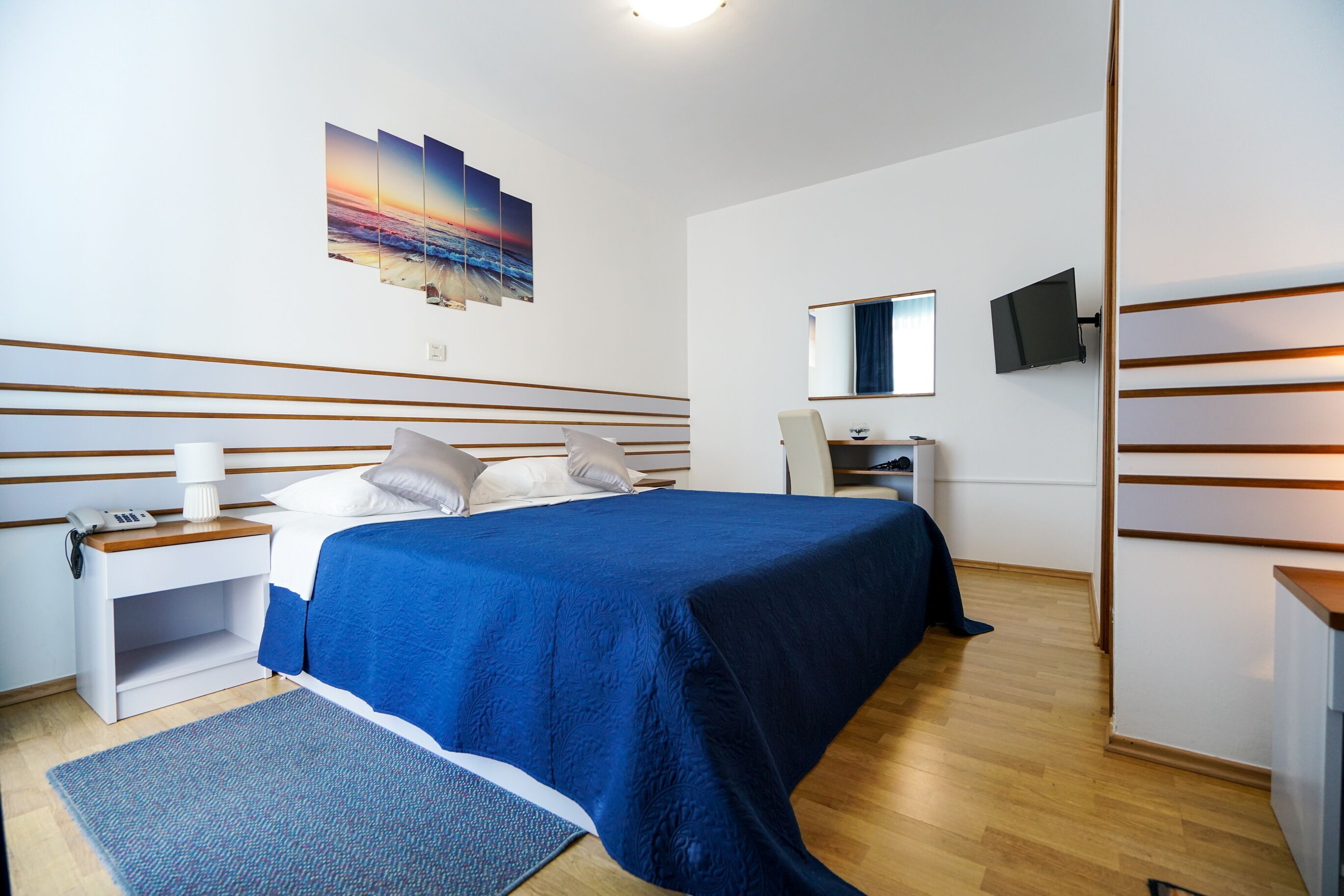 A modern hotel room with a large bed, a bedside table with a lamp, a flat-screen TV, and a painting on the wall.